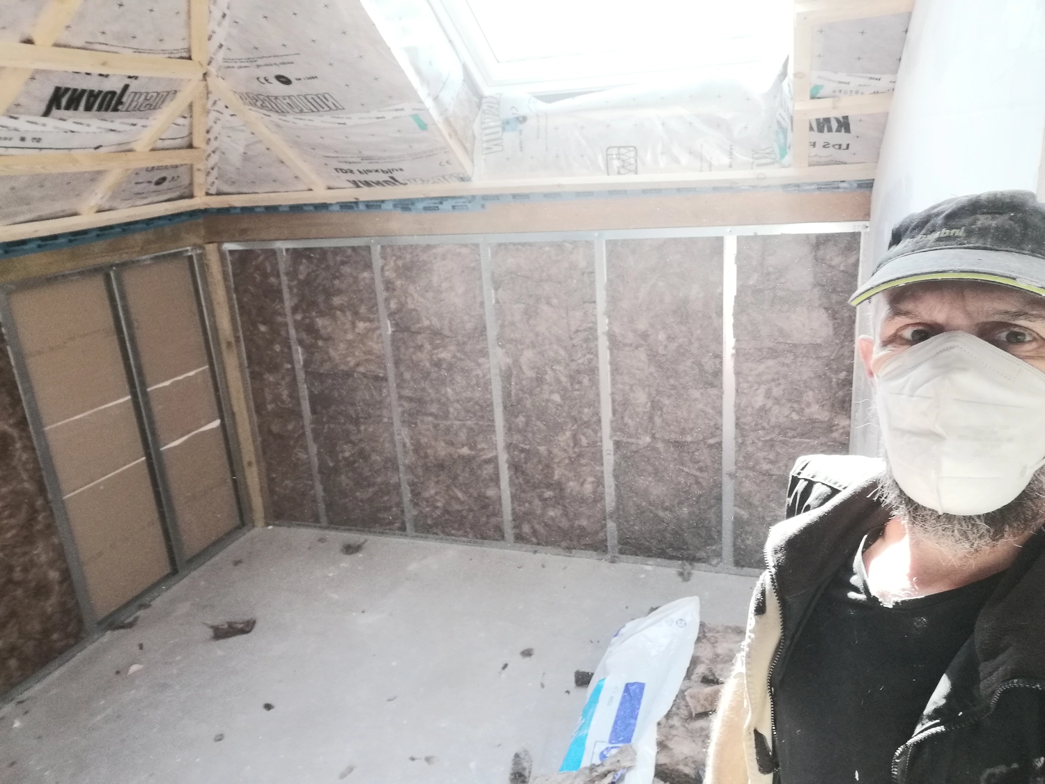 Innenausbau D.Förster - Ein Mann mit Mundschutz und Kappe macht ein Selfie in einem unfertigen Dachbodenzimmer im Bau. Die Wanddämmung und das Fachwerk sind sichtbar.